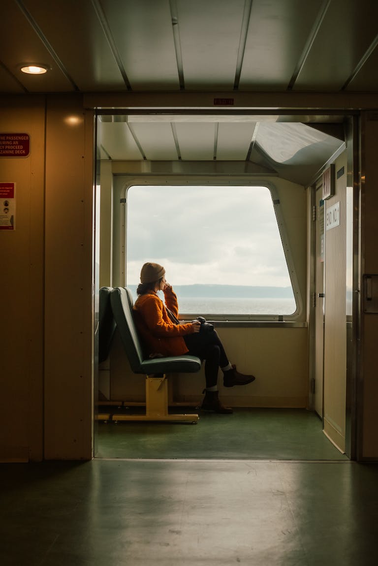 A woman seated in solitude aboard a ferry, gazing at the sea through a window.