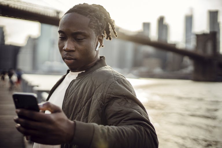 Serious African American man with dreadlocks surfing cellphone while standing on river embankment with bridge and towers on background